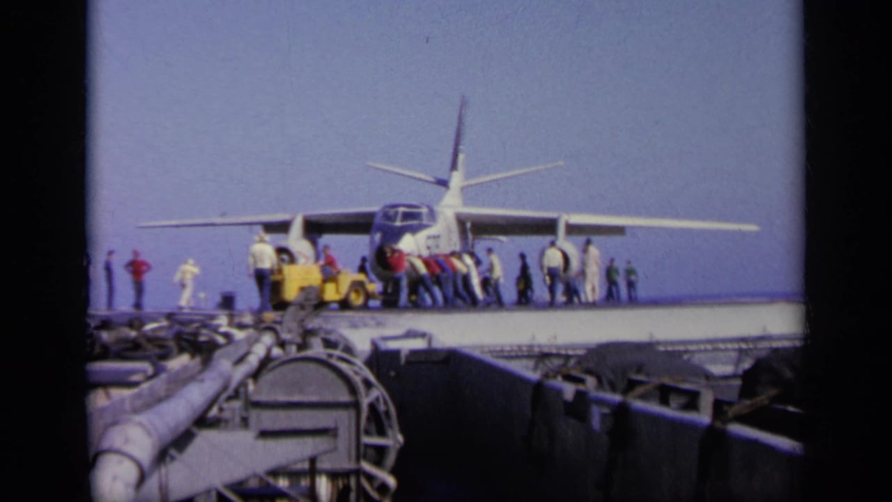 Military jet on a carrier deck with workers and a yellow vehicle nearby.
