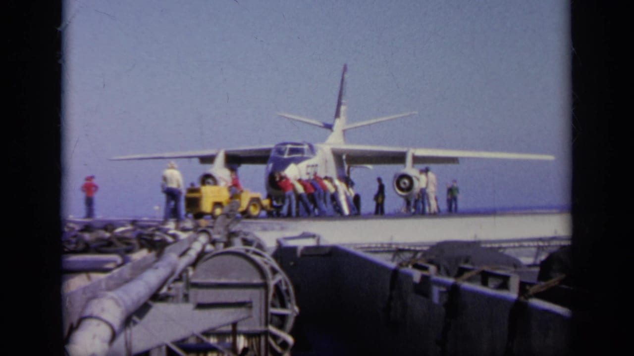 Aircraft facing the camera on a carrier deck as people stand and work nearby.