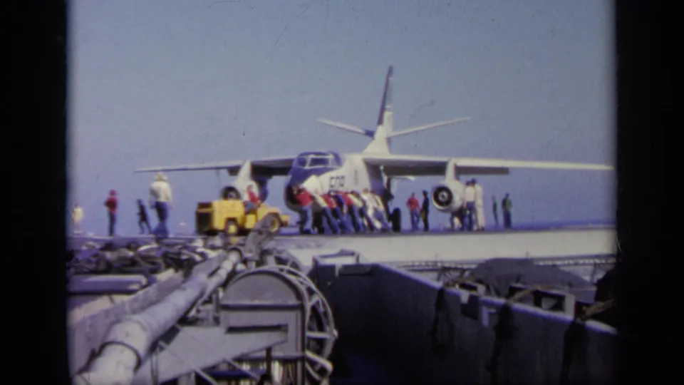 Distant 8mm view of workers pushing or guiding a military jet on a carrier deck, with a yellow service vehicle and ship hardware in front.