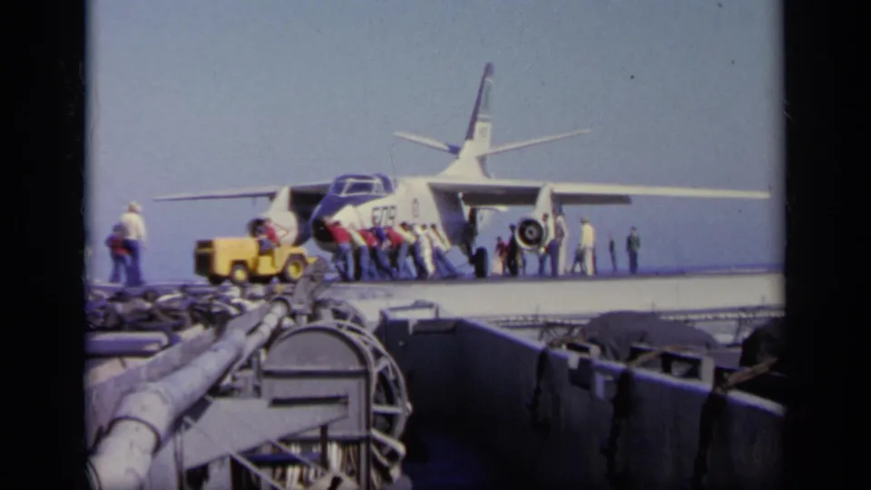 1963 carrier deck frame from Greece metadata, showing a military jet with workers, yellow support vehicle, visible markings, and foreground ship structures.
