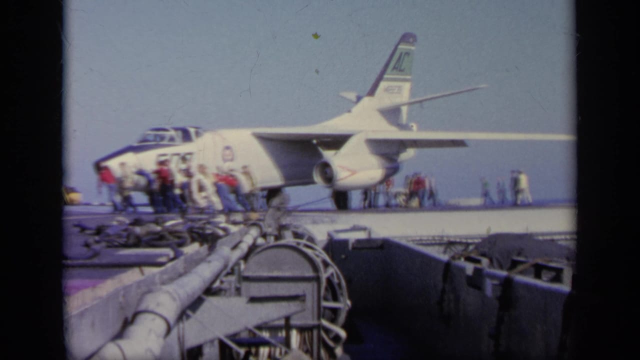 1963 Greece 8mm frame showing a military jet on a carrier deck with crew activity around the nose, wing, and landing gear area.