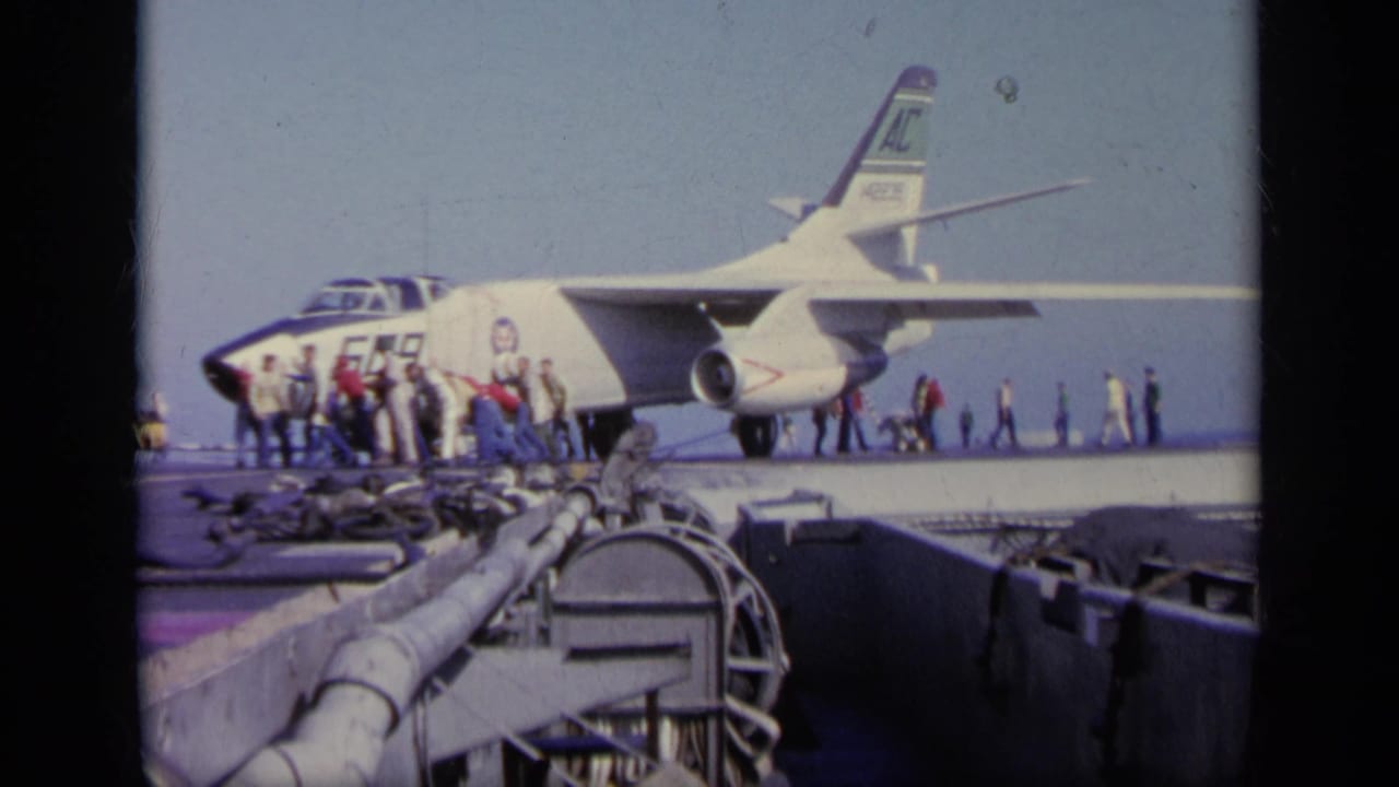 1963 8mm frame of a military jet on an aircraft carrier deck, with sailors and crew visible near the aircraft.