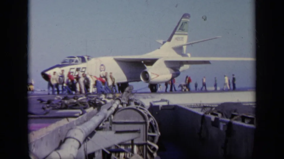 1963 8mm frame of a military jet on an aircraft carrier deck, with sailors and crew visible near the aircraft.