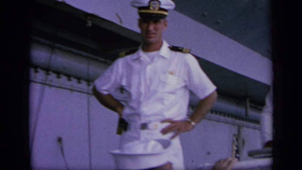 A Navy officer in white dress uniform stands with hands at his sides on a carrier deck, filmed in soft 8mm color.