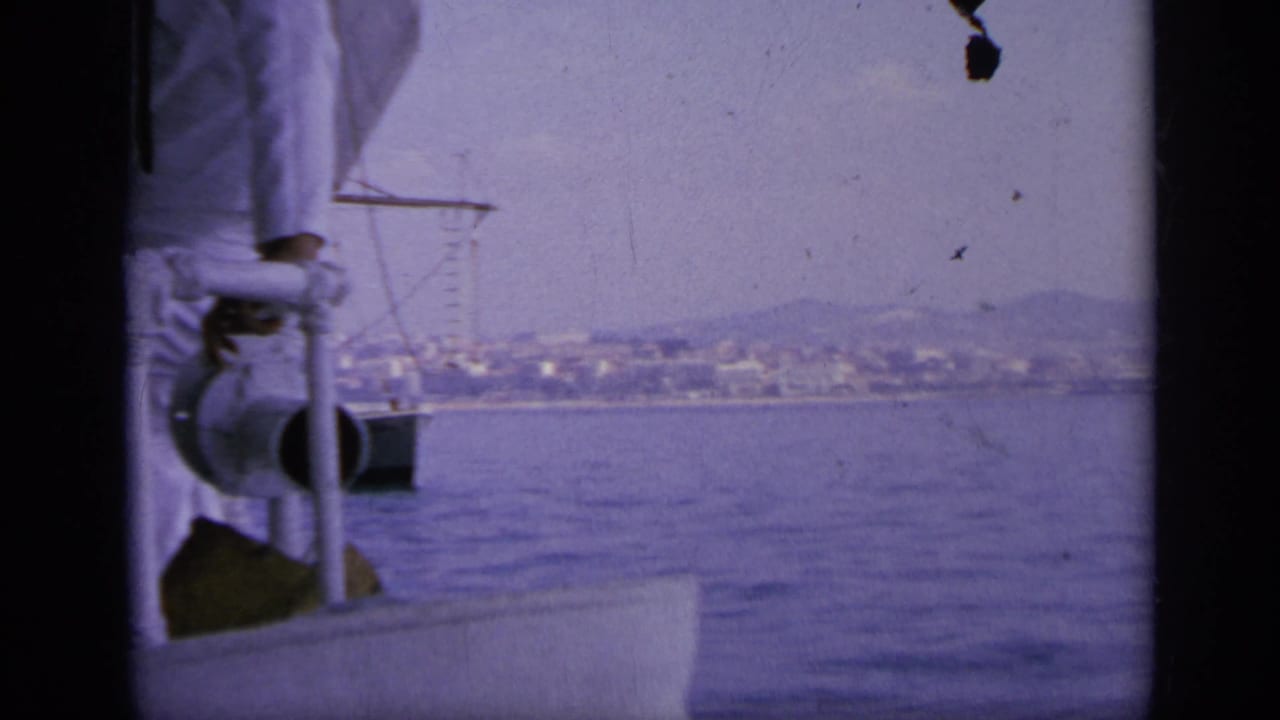 Sailor in white stands by a ship railing as the sea and distant coast appear beyond.
