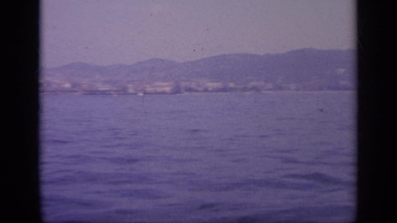 1963 Greece 8mm frame from a ship looking over calm sea toward a soft, mountainous coastline under a pale sky.