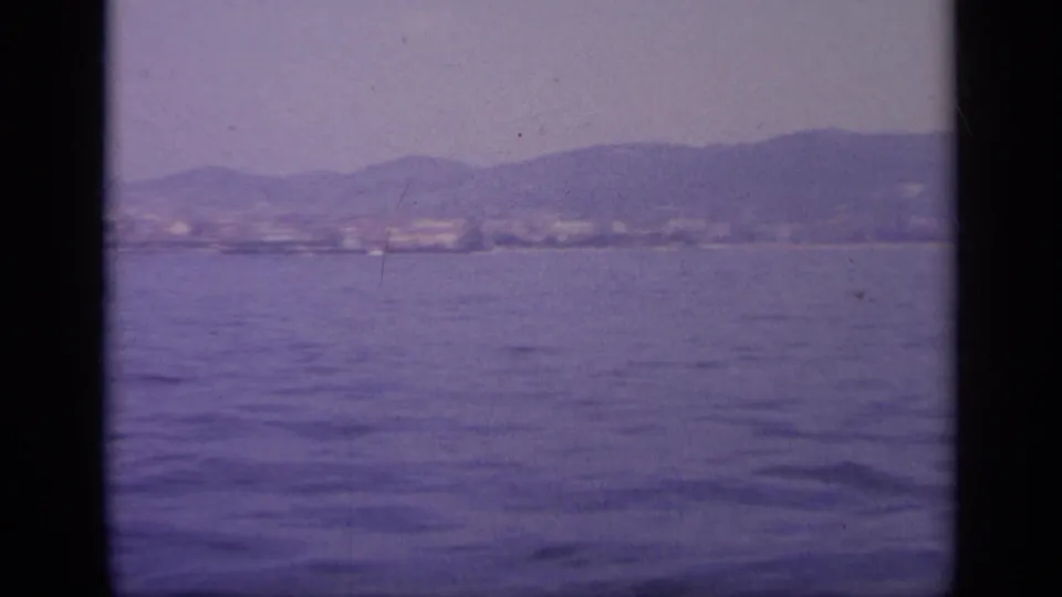 1963 Greece 8mm frame from a ship looking over calm sea toward a soft, mountainous coastline under a pale sky.