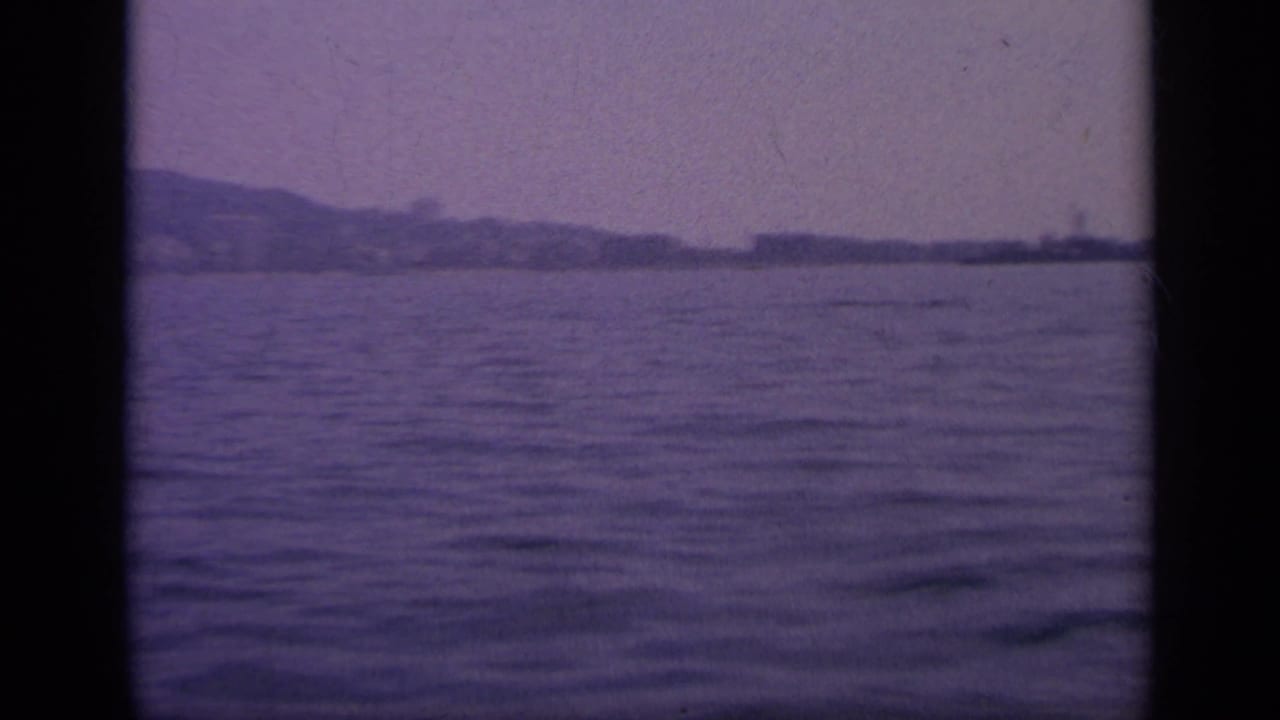 1963 Greece 8mm frame looking over rippled water toward a low, distant shoreline partly obscured by haze and frame borders.