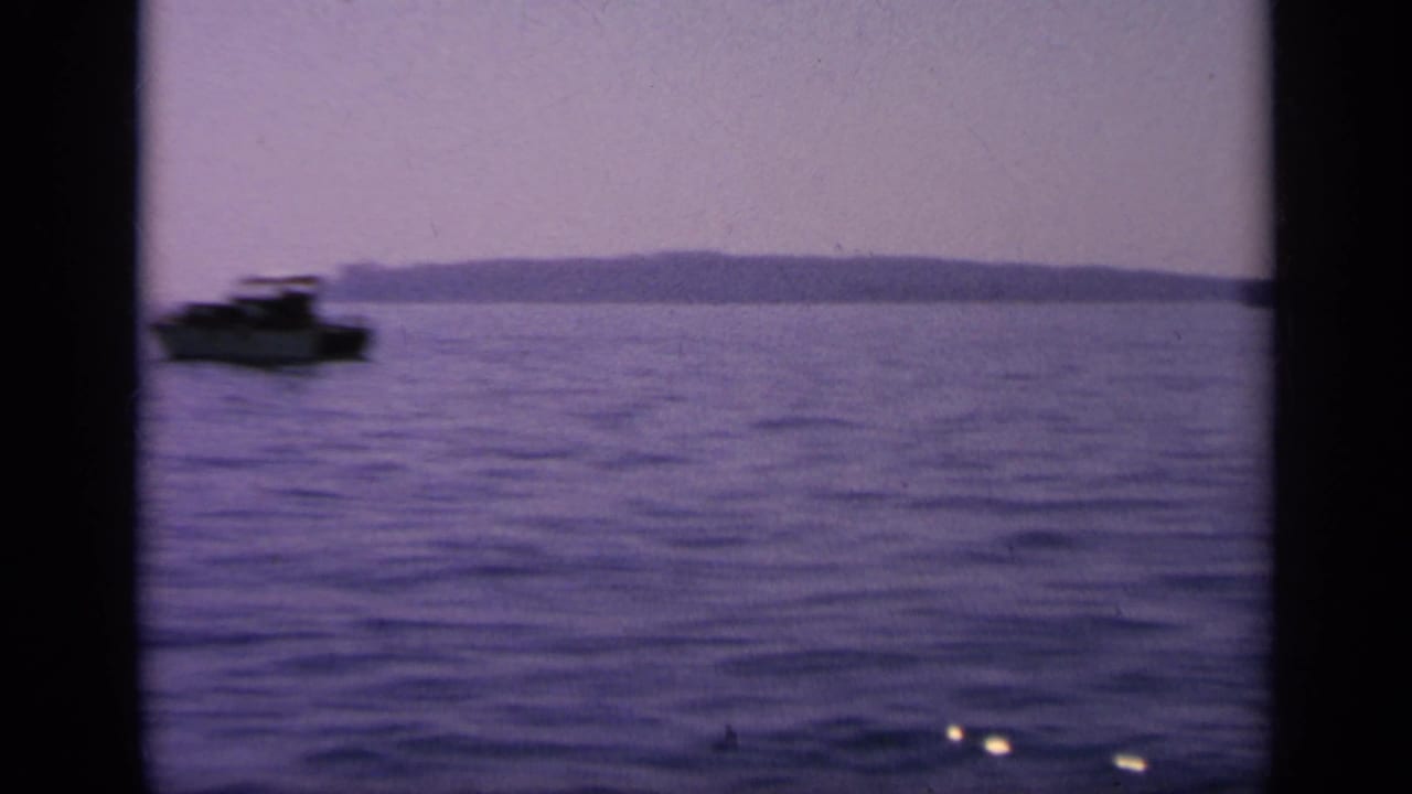 1963 Greece 8mm frame showing a small motorboat moving across calm water with low land on the horizon.