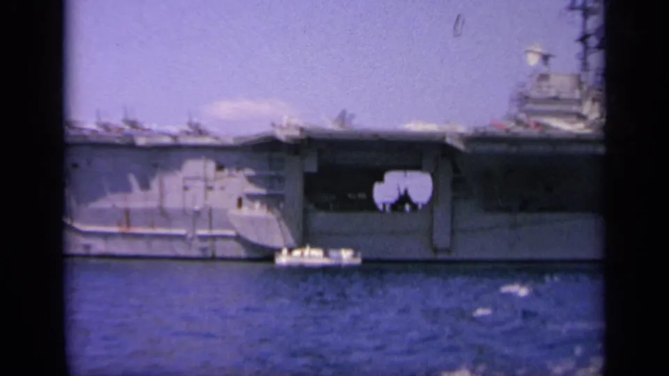 Faded 8mm frame of a U.S. Navy aircraft carrier at sea, showing the midsection, deck edge, and a small boat alongside.