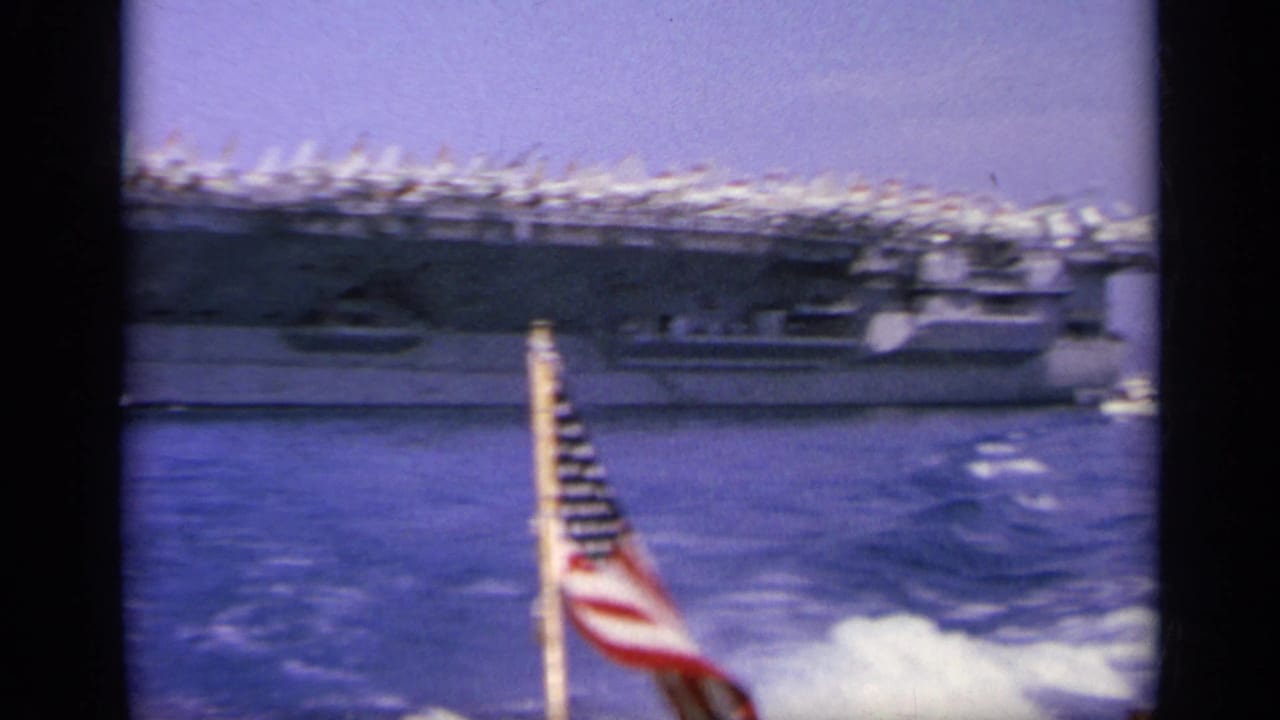 1963 8mm shot near Greece of an aircraft carrier across choppy water, with a U.S. flag visible in the foreground.