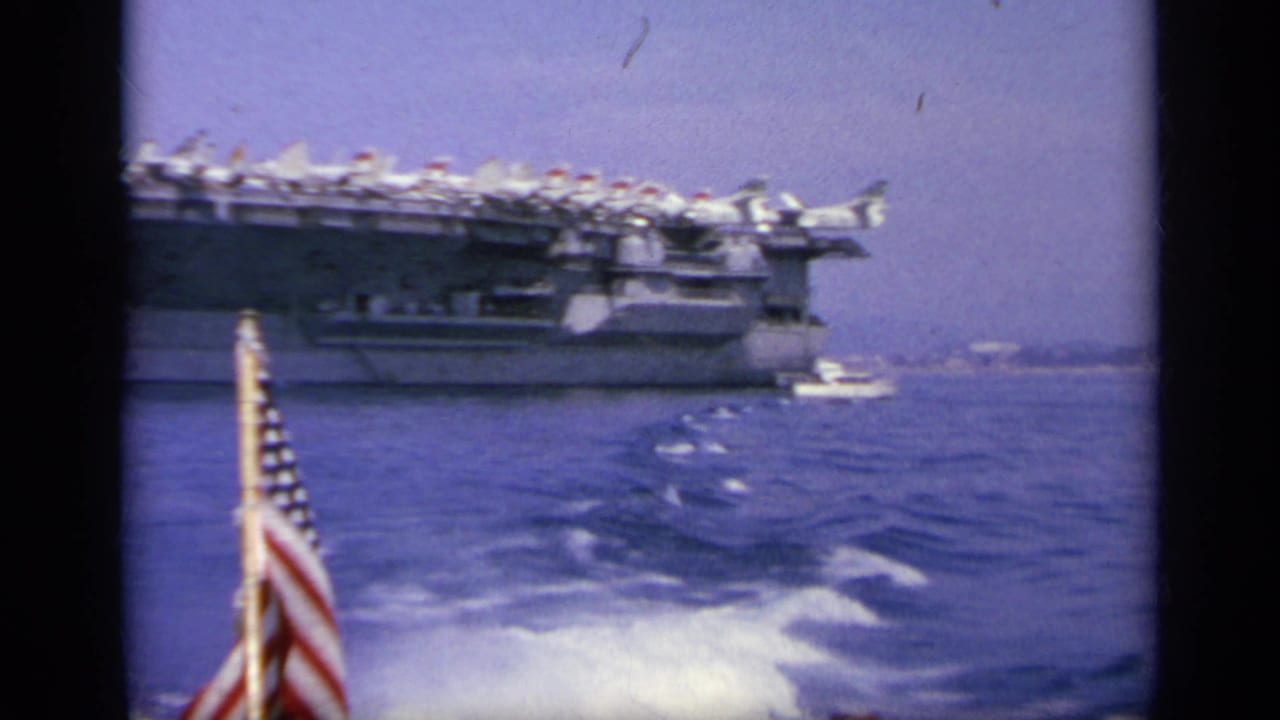 1963 Greece 8mm frame showing a large U.S. Navy aircraft carrier on blue water, with deck jets and an American flag from the filming boat.