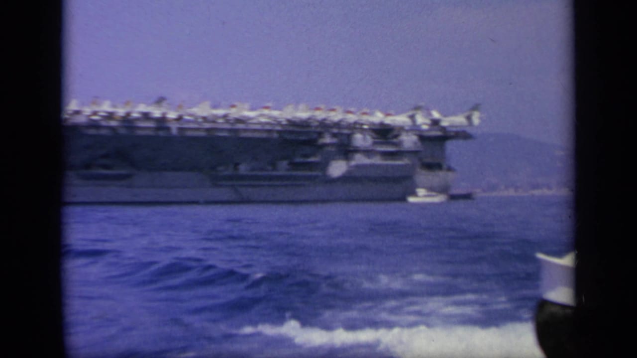 Side view of an aircraft carrier on blue water with small boats nearby.