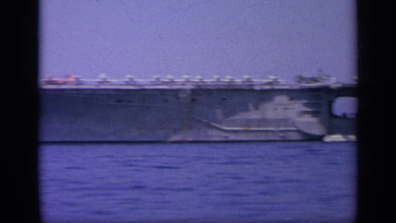 Large aircraft carrier on calm water with mountains in the distance.