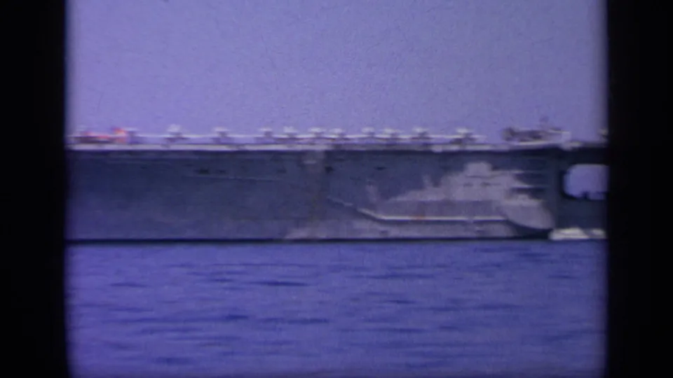 1963 8mm view near Greece showing a naval aircraft carrier at sea, framed by dark film edges and distant mountains.