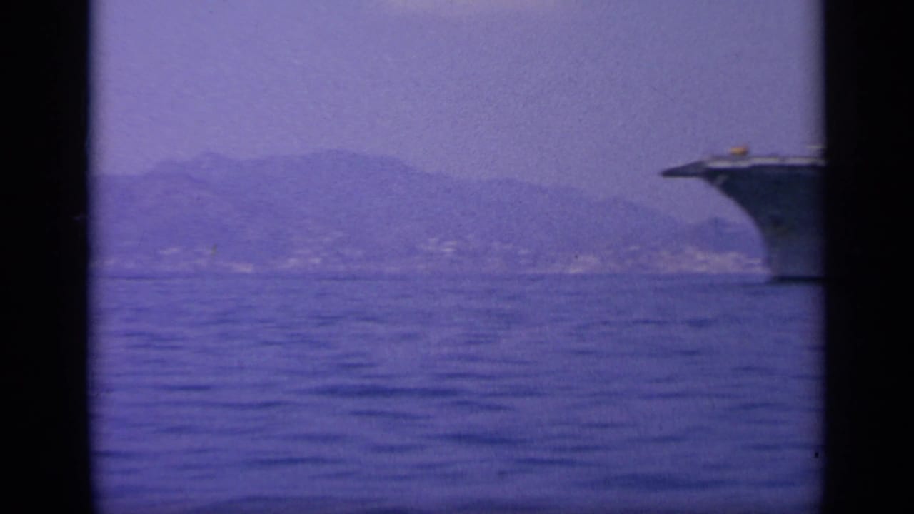 1963 8mm frame near Greece with the bow of a naval carrier at right, open water, and hazy coastal mountains.