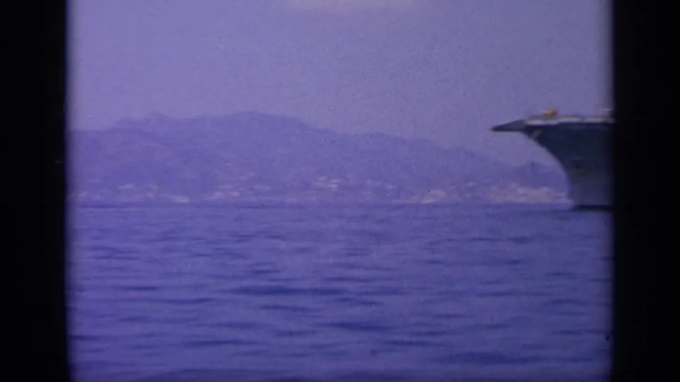 1963 8mm frame near Greece with the bow of a naval carrier at right, open water, and hazy coastal mountains.