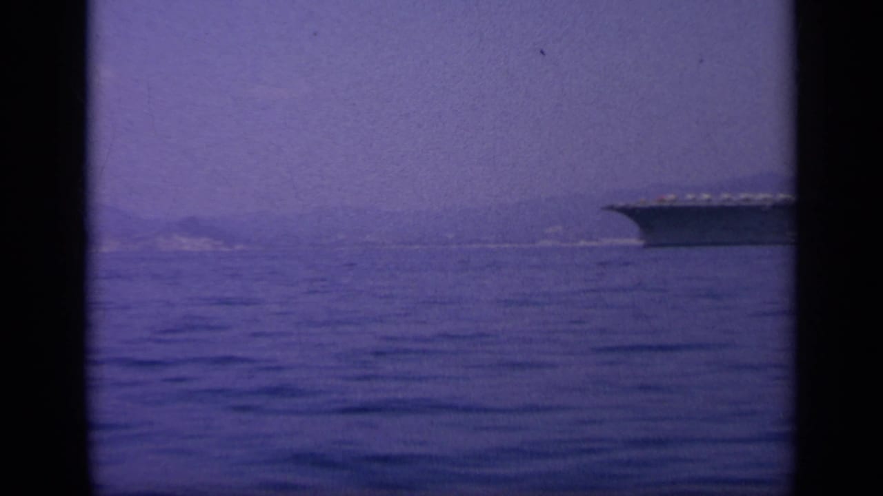 1963 8mm frame near Greece showing the carrier's bow at the edge of a calm sea view with a distant shoreline.
