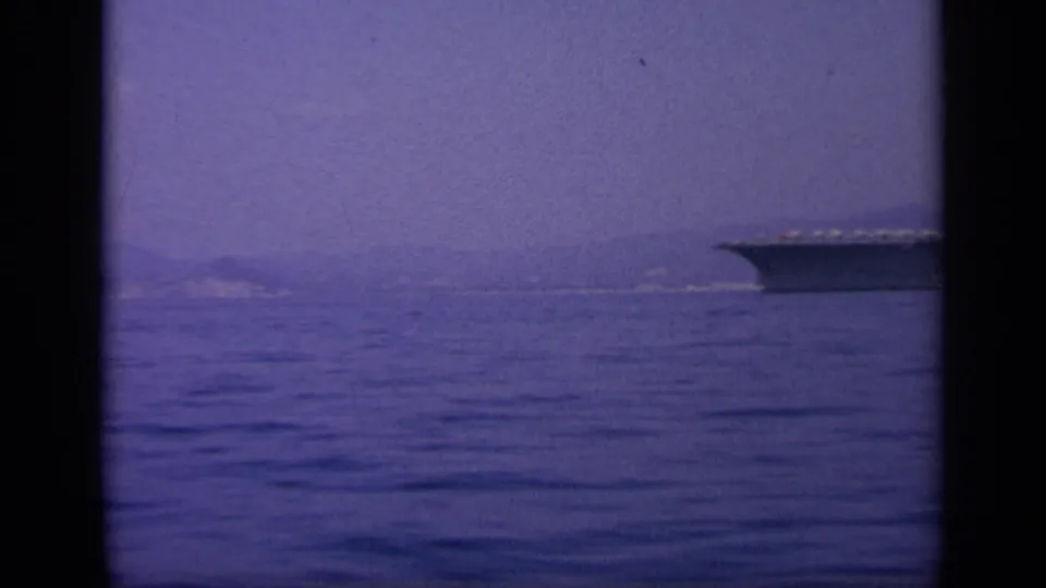 1963 8mm frame near Greece showing the carrier's bow at the edge of a calm sea view with a distant shoreline.