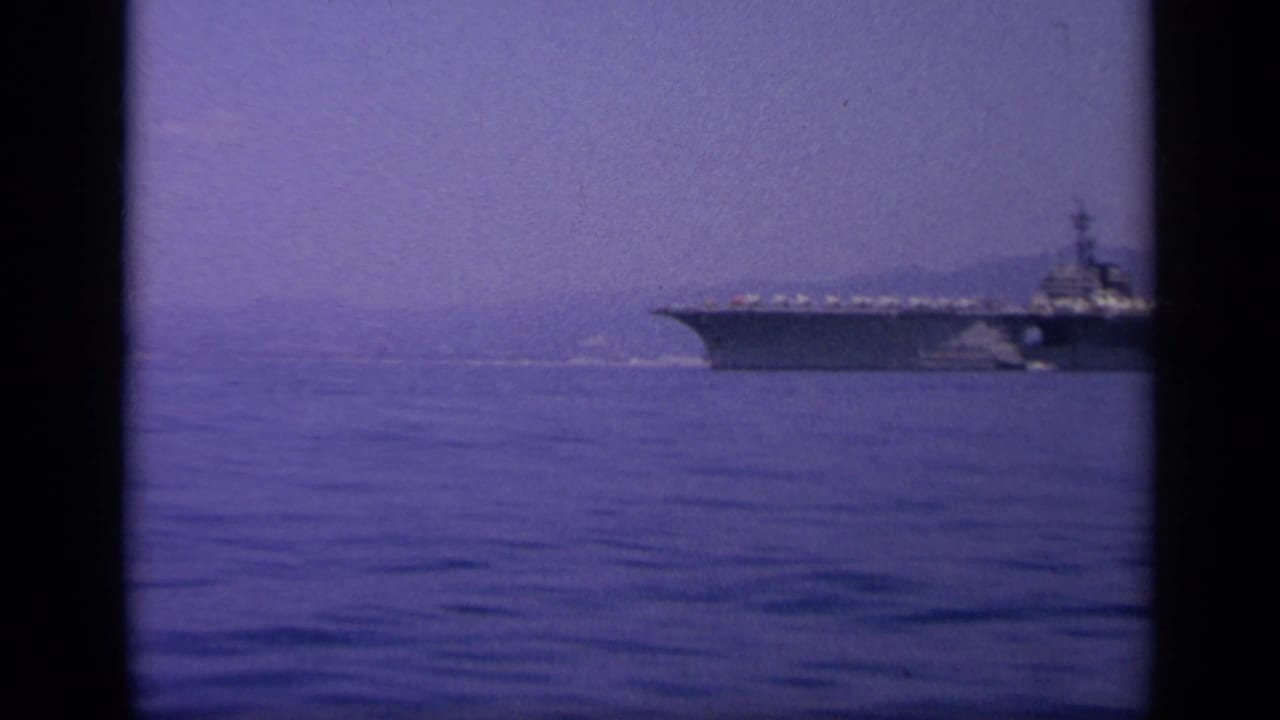 Aircraft carrier on calm water with hazy coastline behind it.