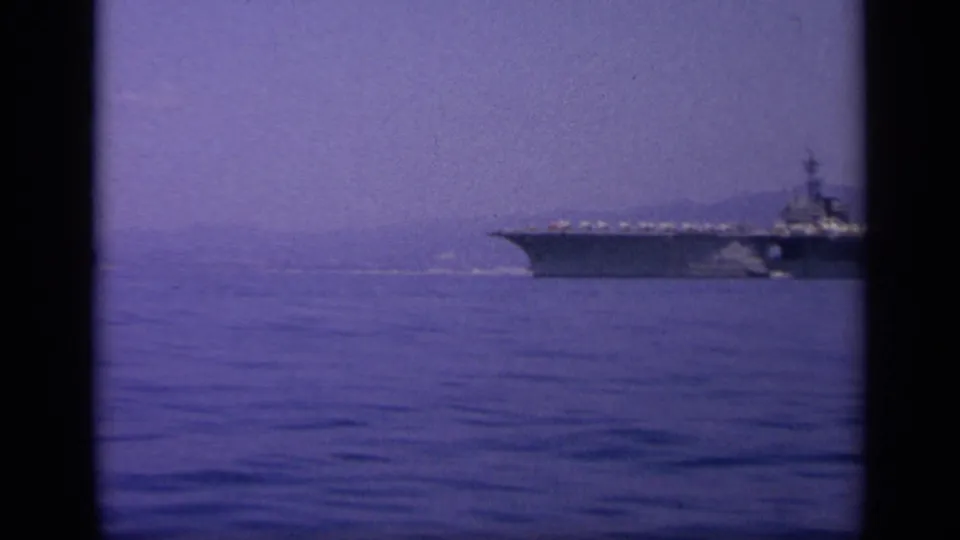 1963 Greece 8mm frame showing an aircraft carrier moving across a calm bay with hills and shoreline in the background.
