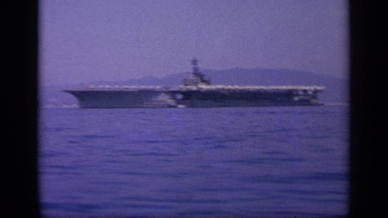 A large aircraft carrier is seen in profile on a calm Greek bay, with a hazy hillside coast behind it.