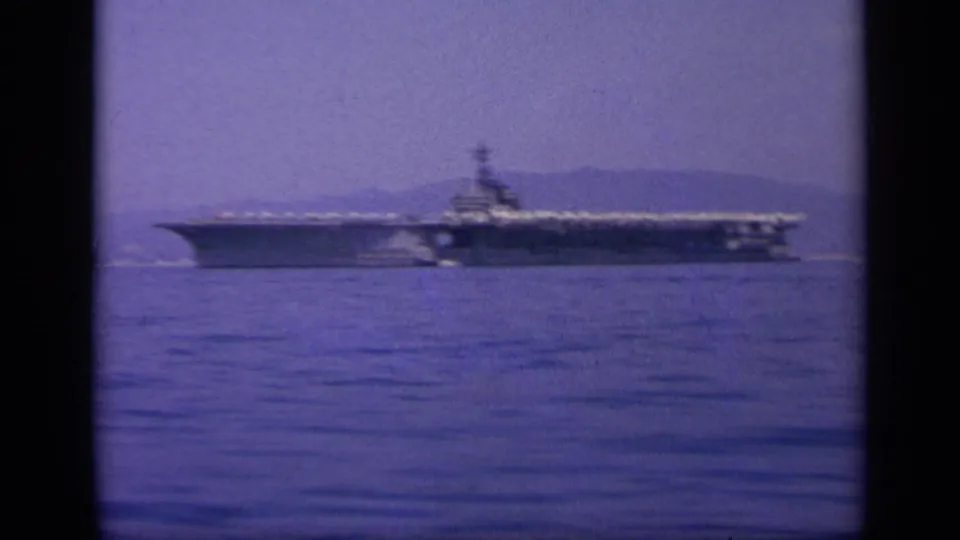 A large aircraft carrier is seen in profile on a calm Greek bay, with a hazy hillside coast behind it.
