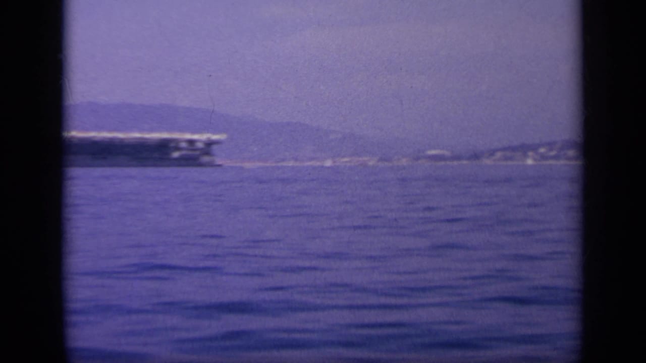 The carrier moves partly out of frame, leaving calm water and a distant coastal hillside visible in the 1963 Greece footage.