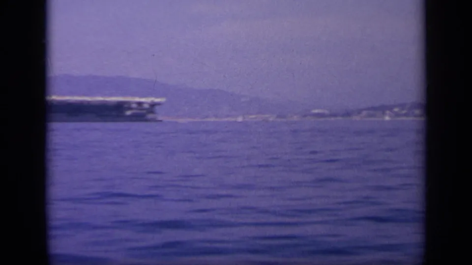 The carrier moves partly out of frame, leaving calm water and a distant coastal hillside visible in the 1963 Greece footage.