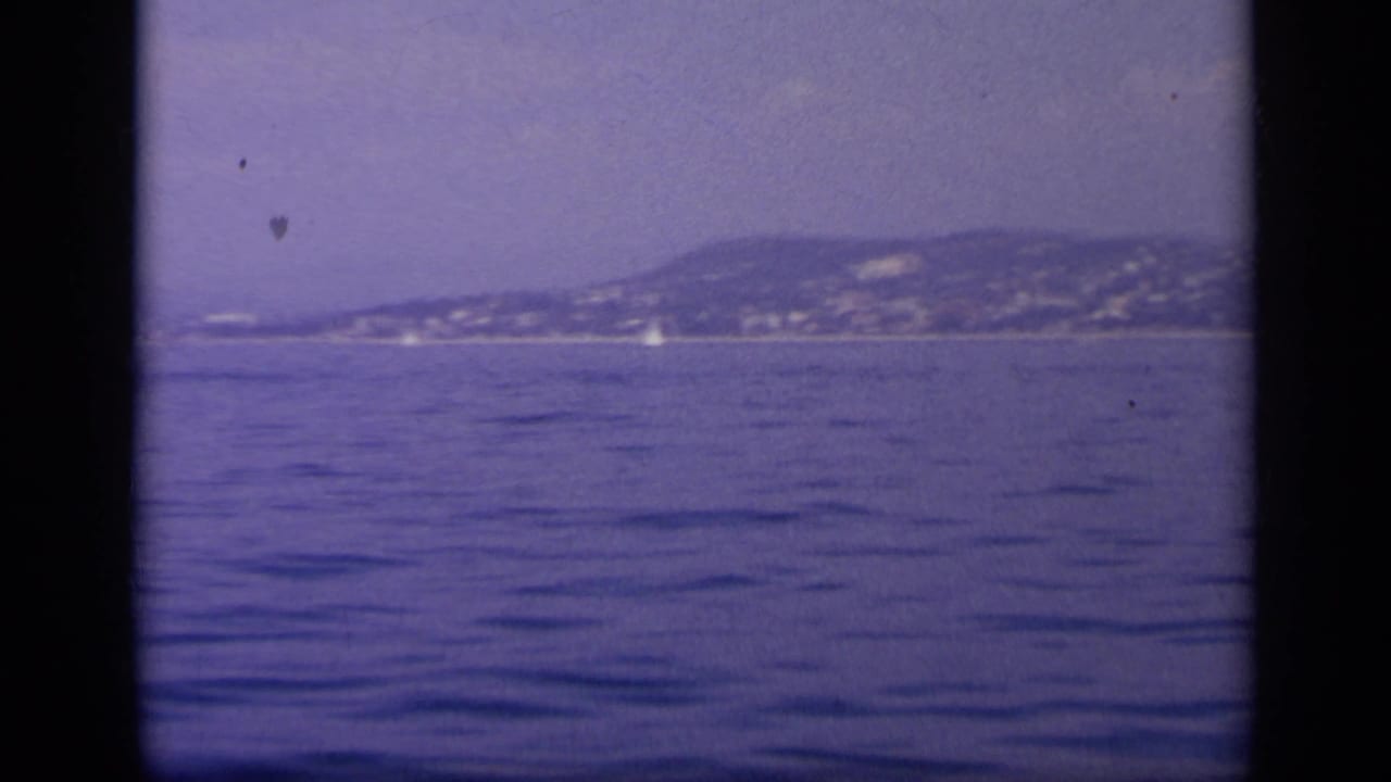 A calm Greek bay fills the frame with a hazy hillside shoreline in the distance after the carrier has moved out of view.