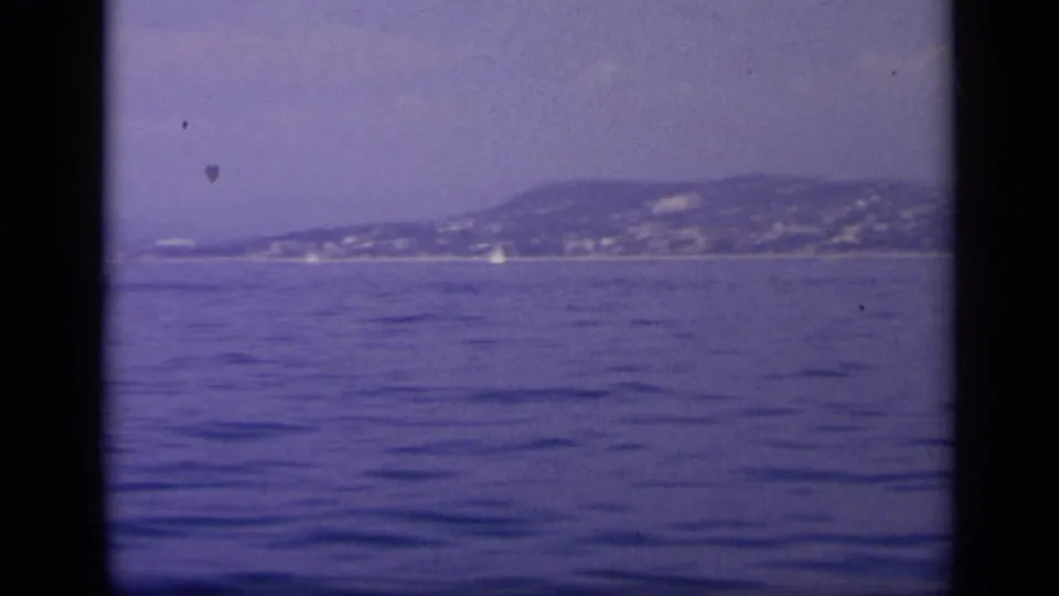 A calm Greek bay fills the frame with a hazy hillside shoreline in the distance after the carrier has moved out of view.