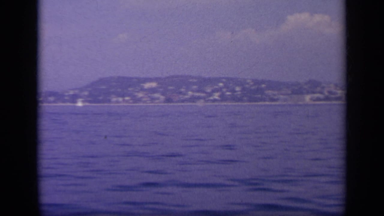 Distant coastal hillside city across calm water under a hazy sky.
