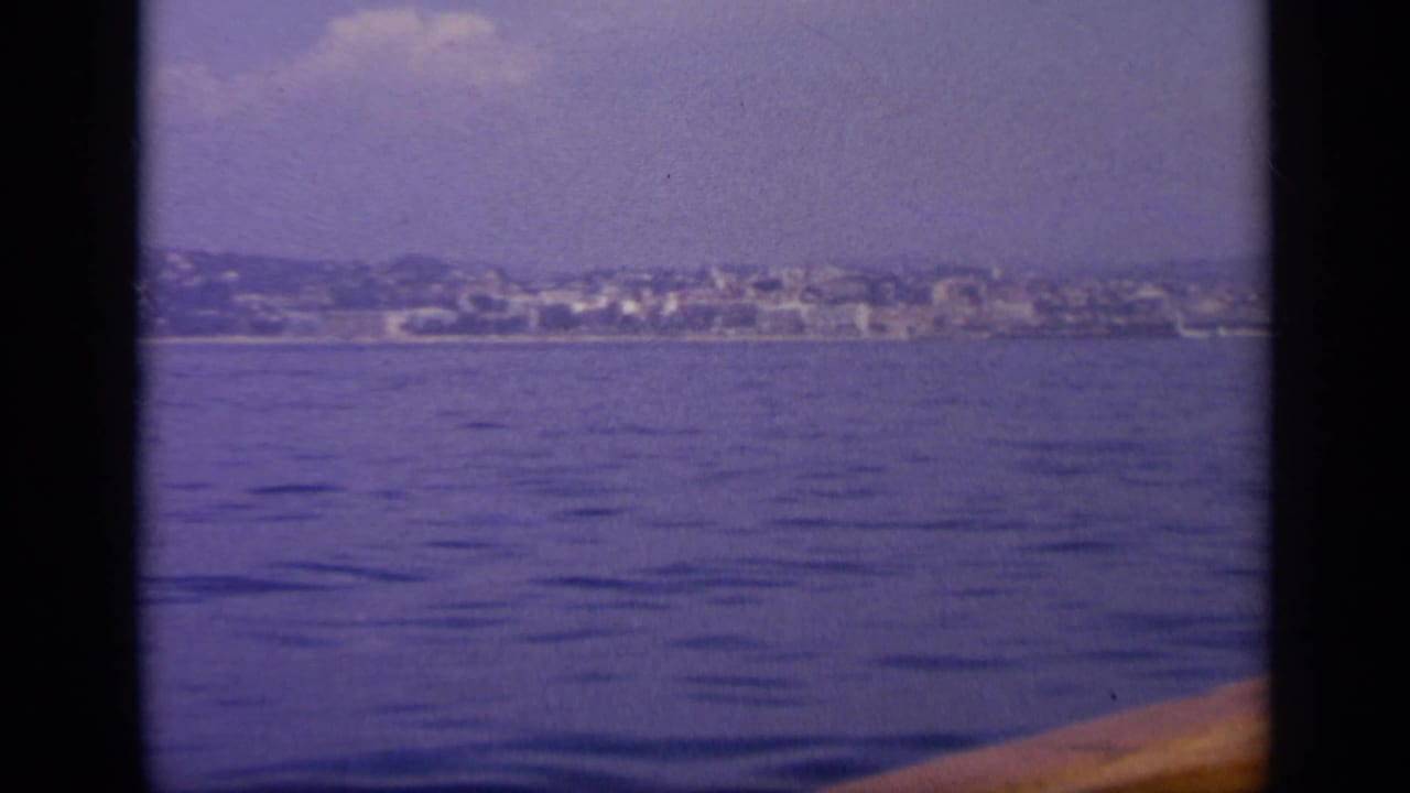 Boat-level 1963 Greece view of a distant coastal town and low hills, partly framed by dark film-gate bars.