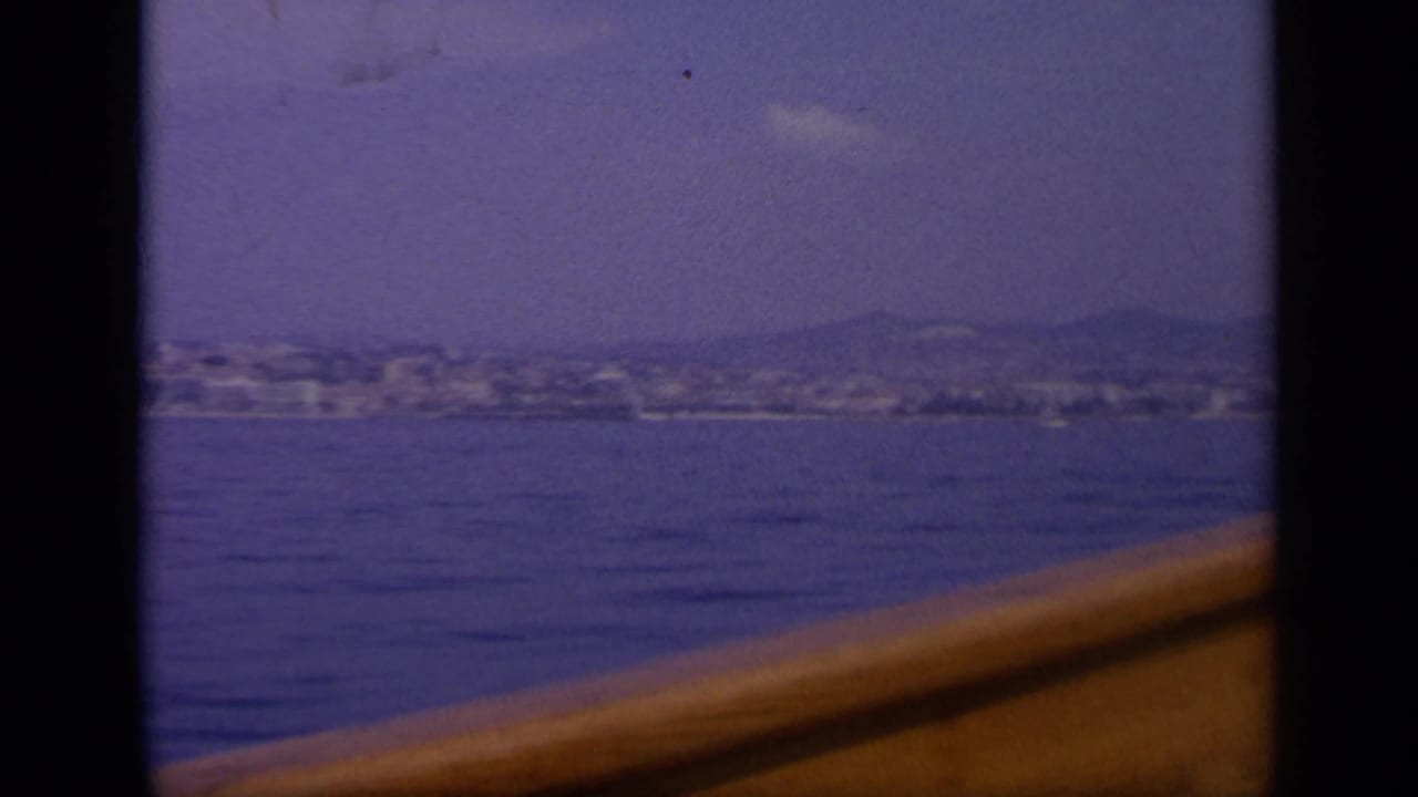 1963 Greece home-movie frame showing a low shoreline city across water with a warm-toned boat rail in front.