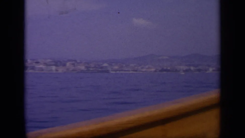 1963 Greece home-movie frame showing a low shoreline city across water with a warm-toned boat rail in front.
