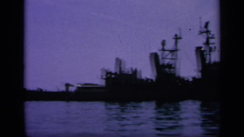 Distant naval ship shapes in a Greek harbor, filmed on 8mm with a purple twilight cast and soft low-light detail.