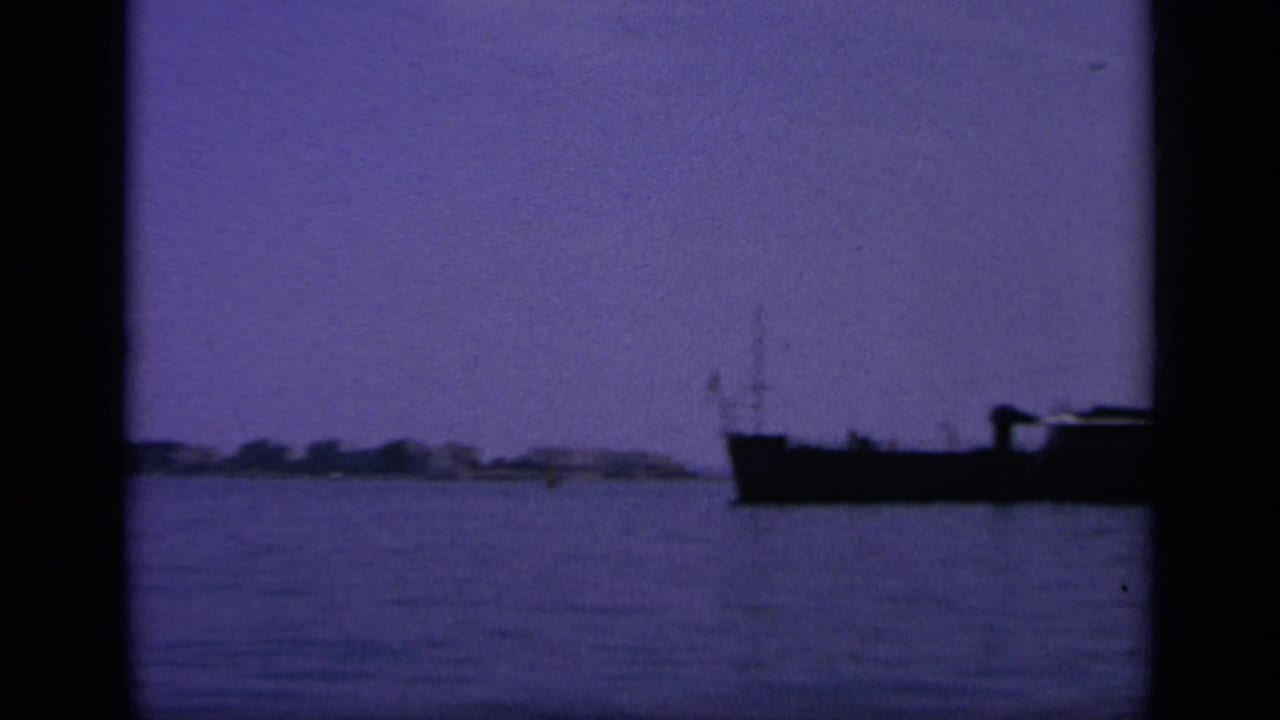 8mm frame from Greece in 1963 showing a low dark boat against open harbor water and a faint distant shore.