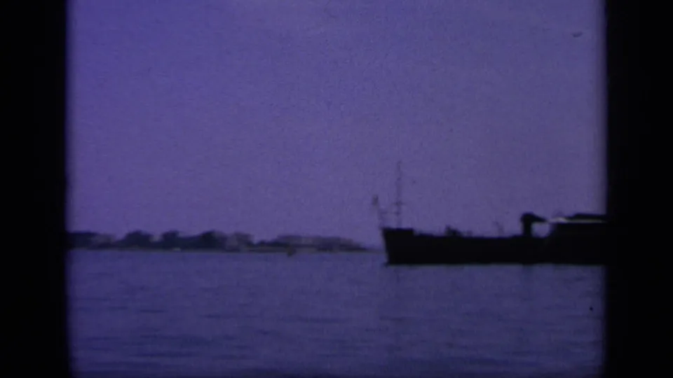 8mm frame from Greece in 1963 showing a low dark boat against open harbor water and a faint distant shore.