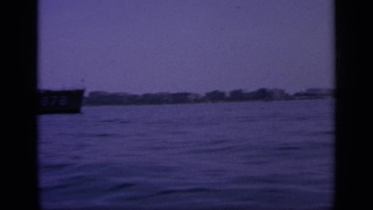 Wide 1963 Greek harbor view on 8mm film, with a partial dark boat at left and low shoreline across the water.