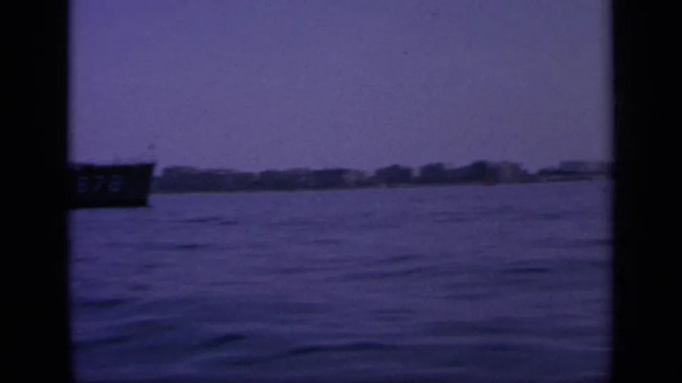 Wide 1963 Greek harbor view on 8mm film, with a partial dark boat at left and low shoreline across the water.