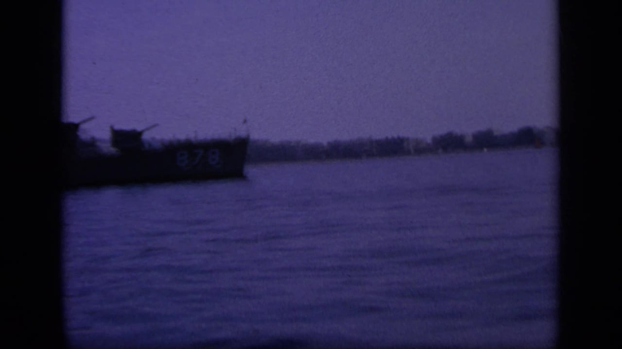 Dark purple-tinted naval ship marked 878 on calm water near a distant shoreline.
