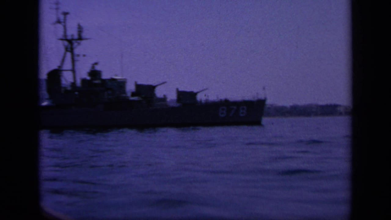 1963 Greece home-movie frame of a naval ship marked 878 moving across a bay, with mast, deck structures, and low shore visible.