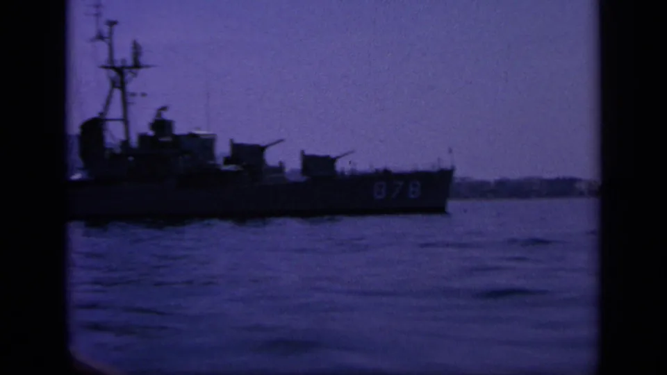 1963 Greece home-movie frame of a naval ship marked 878 moving across a bay, with mast, deck structures, and low shore visible.