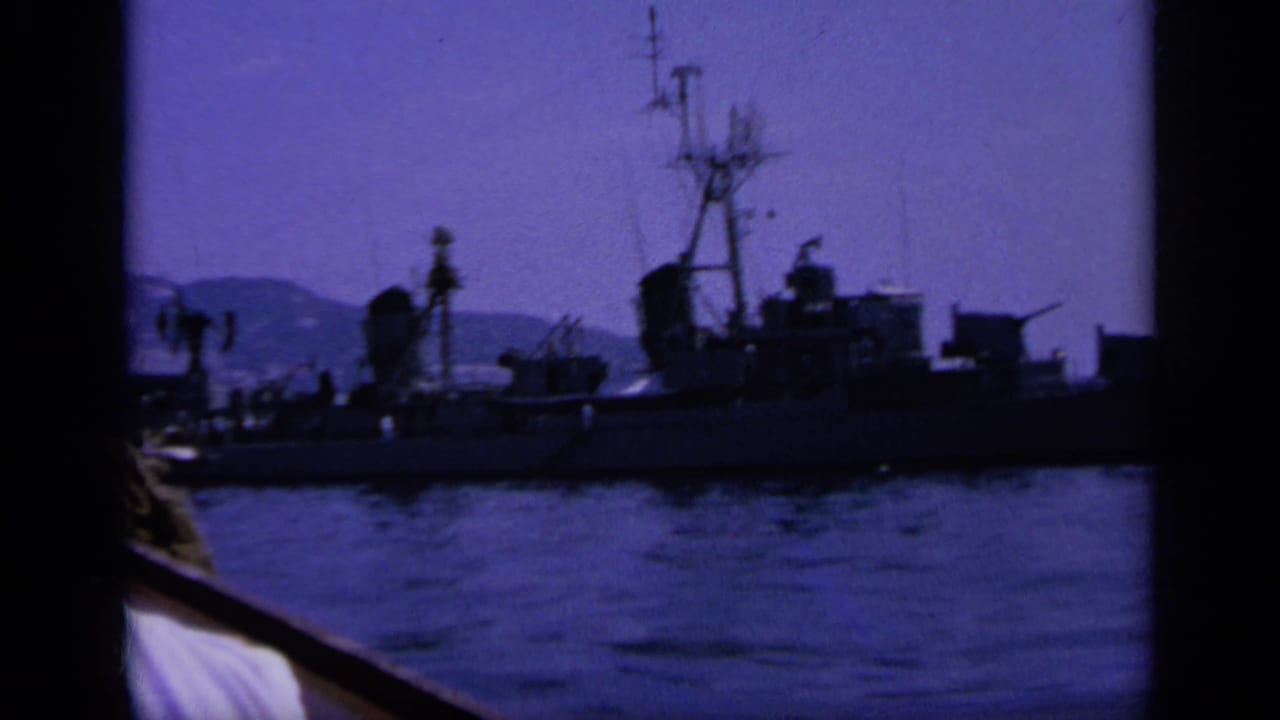 1963 Greece 8mm frame showing a closer naval ship profile, nearby boat rail, bay water, and hazy hills beyond.