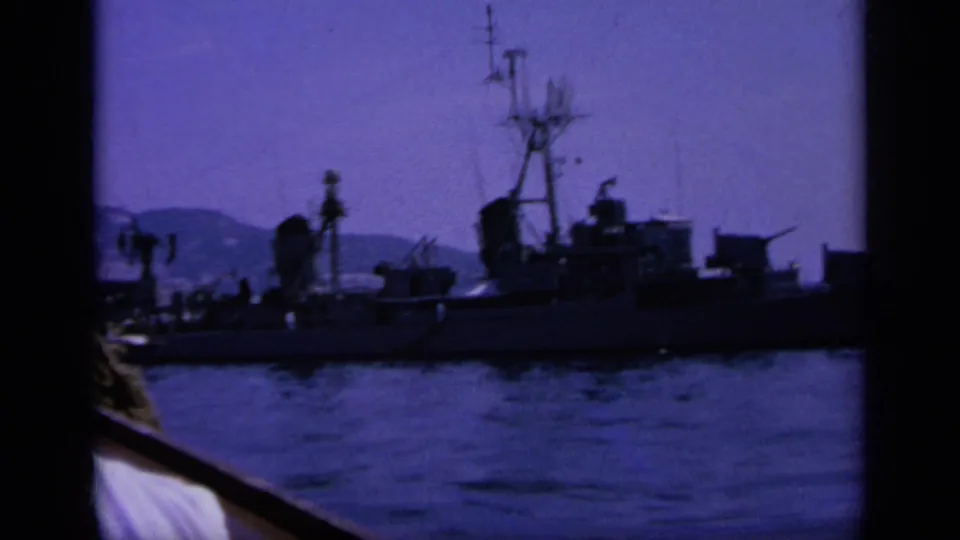 1963 Greece 8mm frame showing a closer naval ship profile, nearby boat rail, bay water, and hazy hills beyond.