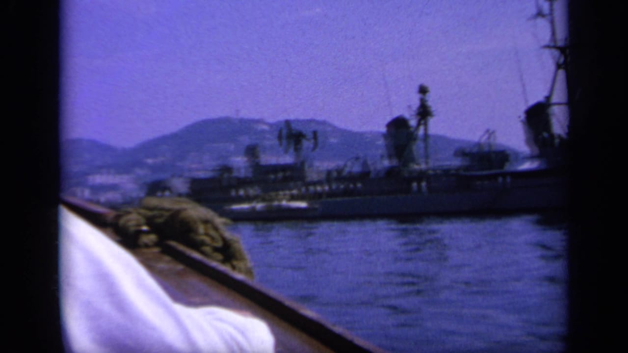 1963 Greece 8mm frame from a small boat looking toward a naval vessel on blue bay water, with hills in the distance.