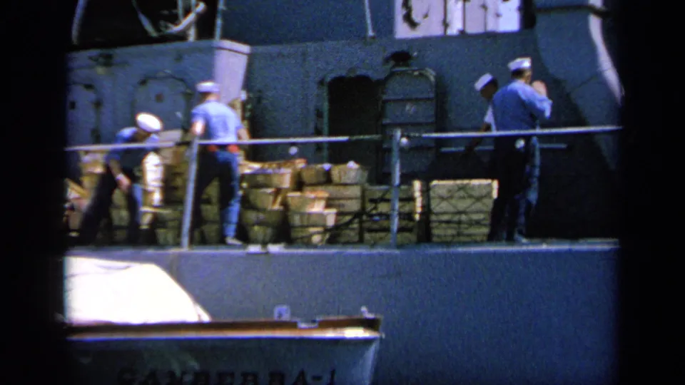 1963 Greece 8mm frame showing sailors on a naval deck with stacked supplies, railings, and a small boat below.