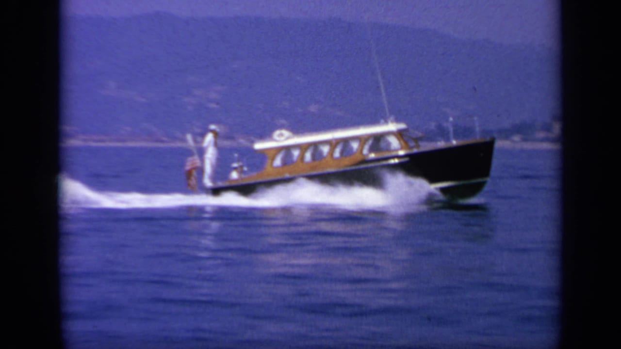 Wooden speedboat crosses blue water with a standing man and small American flag at the stern.