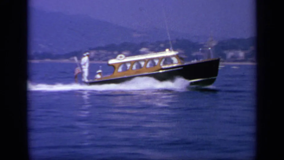 1963 Greece coastal frame showing the wooden speedboat in motion, with passengers aboard and hazy hills beyond the water.