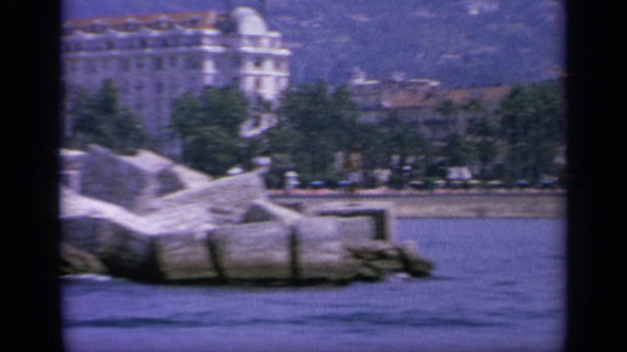 1963 Greece coastal frame with rocks, waterfront trees, and a large pale building seen from the water after the speedboat sequence.